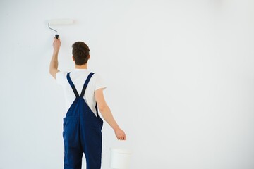 young man in blue overalls painting wall into white color with a roller