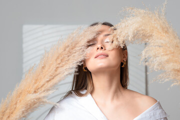 Young beautiful brunette woman in white shirt, stylish and minimalistic portrait in light and beige colors, decorative dried flowers in hand
