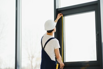 Workman in overalls installing or adjusting plastic windows in the living room at home