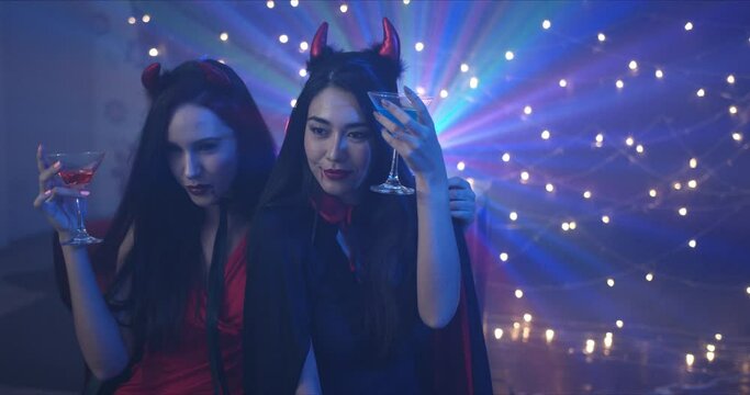 Slow Motion, Handheld - Two beautiful seductive young Caucasian female friends, dressing in she devils drinking and dancing at a Halloween costume party in a dark room with decorative lights.