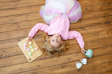 Little beautiful child girl lying on wooden floor with mugs and sweet marshmallows