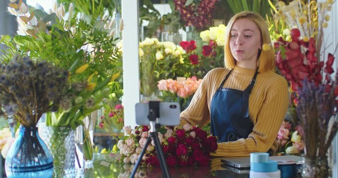 Medium shot of beautiful woman blogger in apron shooting video in flower store with smartphone. Female florist making live conference for followers, viewers. Vlogger telling about flowers in shop.