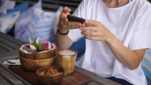 Close up of female hands taking picture of a smoothie served in coconut bowl with granola on a side and coffee. 