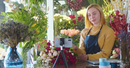 Medium shot of pretty woman blogger in apron shooting video in flower store using phone. Female florist making live conference for followers, viewers. Pretty vlogger telling about flowers in shop.