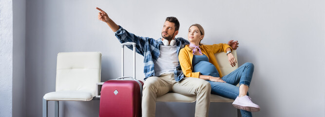 Man pointing with finger near pregnant wife and suitcase in airport, banner