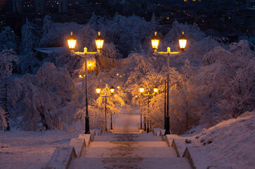 Beautiful view of the night winter city park. A ladder in the snow goes down. Lanterns by the road...