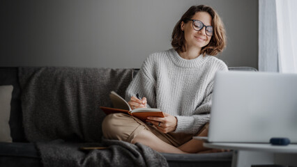 Beautiful young girl student studying at home online while sitting in warm sweater on the couch