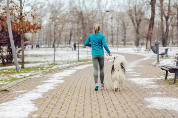 Rear view of sportswoman running together with her dog in park on snowy weather. Winter fitness, pets, friendship, dogs