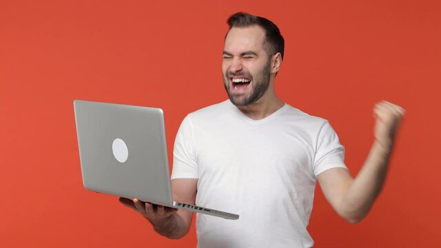 Excited happy bearded young man 20s in white casual t-shirt isolated on orange color background studio. People lifestyle concept. Working on laptop pc computer doing winner gesture screaming say yes