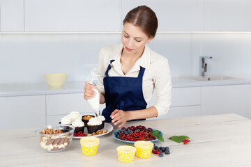 Cute female baker in blue apron  decorating tasty cupcake with creamy top at kitchen table