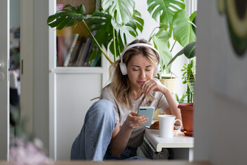 Fototapeta premium Woman listening to music, wear wireless white headphones, using mobile smart phone, chatting in social networks, sitting next to the window, houseplants on windowsill. Life at home. Time to relax. 