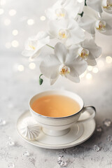 Cozy home spring breakfast still life. A porcelain cup of hot green tea with a marshmallow dessert on a gray background is decorated with a flowering branch of a white otchid. Spring concept. 