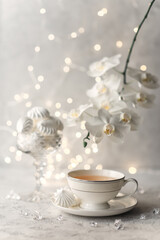 Cozy home spring breakfast still life. A porcelain cup of hot green tea with a marshmallow dessert on a gray background is decorated with a flowering branch of a white otchid. Spring concept. 
