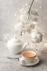 Cozy home spring breakfast still life. A porcelain cup of hot green tea with a marshmallow dessert on a gray background is decorated with a flowering branch of a white otchid. Spring concept. 