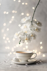 Cozy home spring breakfast still life. A porcelain cup of hot green tea with a marshmallow dessert on a gray background is decorated with a flowering branch of a white otchid. Spring concept. 