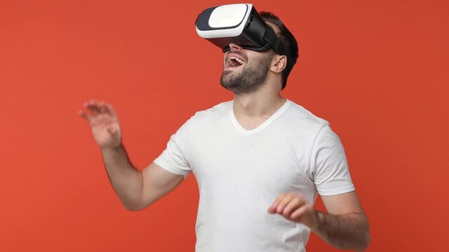 Shocked excited man in white t-shirt isolated on orange background . People lifestyle concept. Watching in vr headset gadget touch something like push click on button point at floating virtual screen