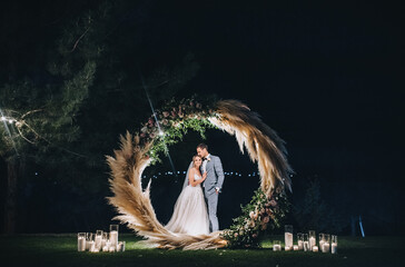 A young groom in a gray suit and a cute, smiling blonde bride embrace, standing on the green grass, near a round wooden arch decorated with reeds, flowers, candles. Night wedding portrait.