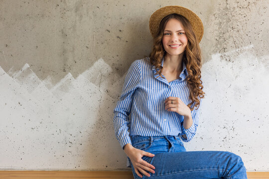 young attractive stylish woman sitting at wall background, straw hat