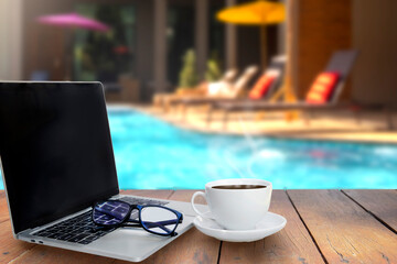 work space  with coffee cup ,, eye glasses and laptop on a wooden table on swimming pool in resort  background.