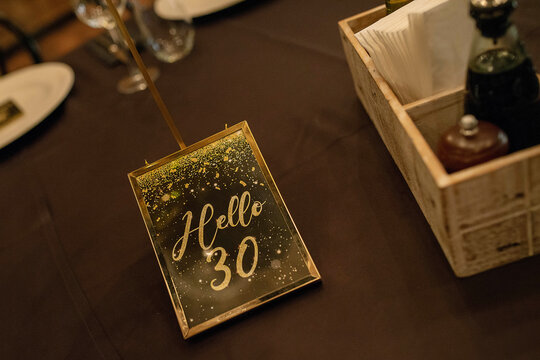 Invitation in gold and black to a 30th years birthday party framed in gold that reads "Hello 30" set on a table, elegant decoration at a restaurant prepared for the celebration of somebody turning 30
