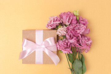Gift box with a tied bow and bouquet of eustoma flowers on a golden background. Minimalistic festive composition.