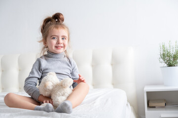 little girl in grey turtleneck playing with teddy bear on white bedding