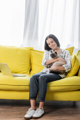 smiling freelancer sitting on sofa near laptop and hugging fluffy cat