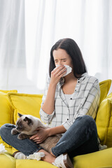 young woman sneezing in paper napkin while sitting on couch with cat and suffering from allergy