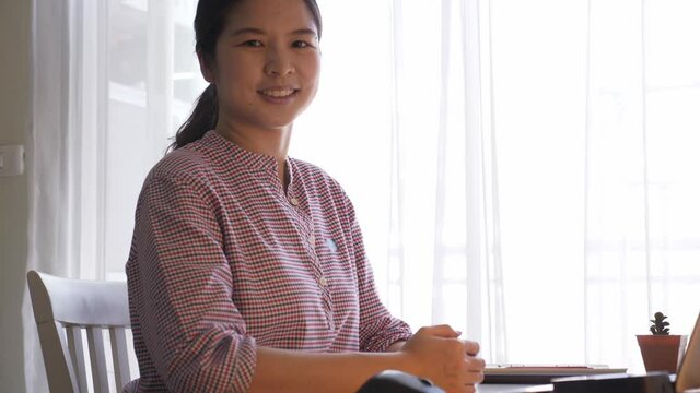 Portrait, handheld, side view, low angle shot of young beautiful Asian woman working from home with laptop, turing face and looking at camera, smiling and waving hand.