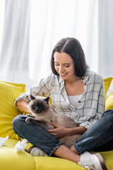 happy woman sitting on sofa with crossed legs and cuddling cat