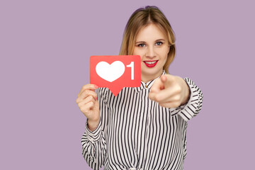 Hey you, put like to content! Pretty woman with red lips in glamour blouse holding social media heart icon , pointing to camera, recommending to follow blog. studio shot isolated on purple background