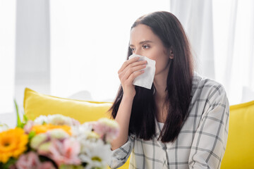 young woman wiping nose while suffering from allergy near flowers on blurred foreground