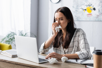 Obraz premium allergic woman wiping nose with paper napkin while working at laptop on blurred foreground