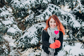 A girl with red hair holds a heart on a stick in her hands against a background of trees and snow. Valentine's day, winter, love, feelings, discounts and promotions.