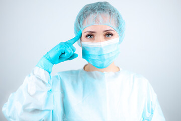 a nurse in a medical gown, mask and protective gloves points a finger at her head