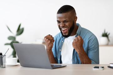 Joyful Black Guy With Laptop Raising Fists At Workplace, Celebrating Business Success