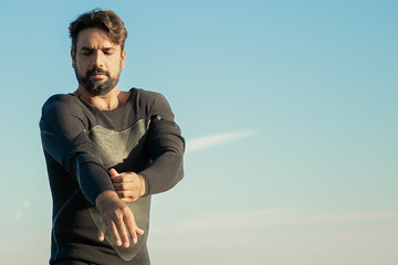 Focused sportsman putting on wetsuit for surfing on ocean beach. Low angle, blue sky background. Active lifestyle concept