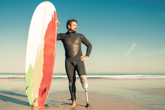 Confident Handicapped Man Standing On Sea Beach With Board. Attractive Brunette Man With Artificial Leg Wearing Black Wetsuit And Looking At Ocean. Physical Disability And Extreme Sport Concept