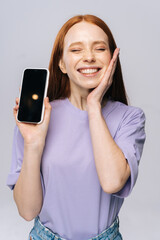 Close-up of happy young woman in casual clothes showing phone with black empty mobile screen, holding hand near face. Pretty lady model with red hair emotionally showing facial expressions in studio.