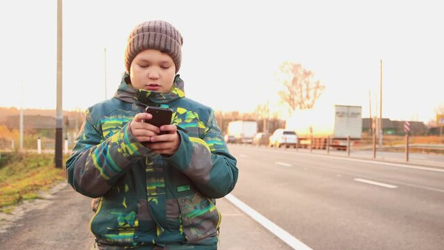Against the background of the highway, a boy is talking on the phone online. Out of focus cars and trucks zip by. Walk down the street in autumn. At sunset, in the evening, a warm soft light.