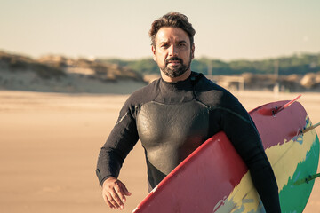 Attractive brunette surfer walking and sand beach with board. Caucasian bearded man holding surfboard and wearing wetsuit. Blurred background. Lifestyle, vacation and summer activity concept