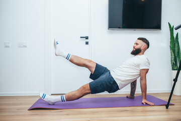 Naklejka premium Young man making sport fitness exercises at home. Training during quarantine. Online training in front of the smartphone camera.