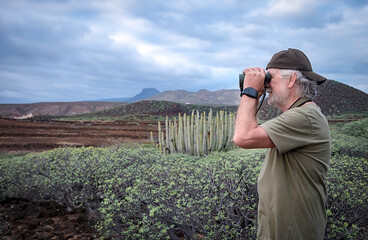 Side view of senior man with white beard looking at the horizon with binoculars. Active retiree enjoying the hike in nature