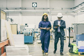 Serious male inspector and black female factory employee in hardhats walking on plant floor and talking, man using tablet. Front view, full length. Labor and production concept
