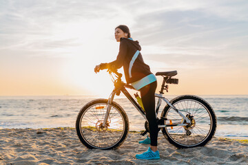 young attractive slim woman riding bicycle, sport on morning sunrise summer beach in sports fitness...