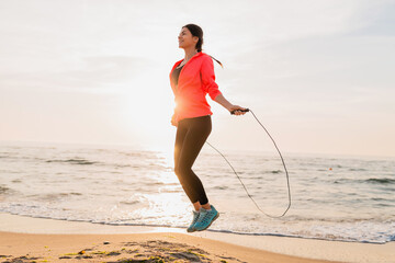 young attractive woman doing sport exercises in morning sunrise on sea beach in sports wear, healthy lifestyle, listening to music on earphones, wearing pink windbreaker jacket, jumping in jump rope