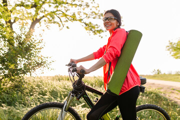 slim fit beautiful woman doing sports in morning in park riding on bicycle with yoga mat in...