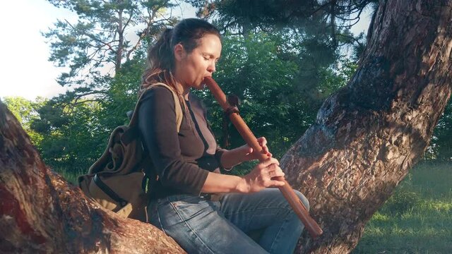 beautiful girl sits on a tree and plays a pimak pipe in a park against the background of nature. Pimak wind instrument Native American Indian Flute of Love
