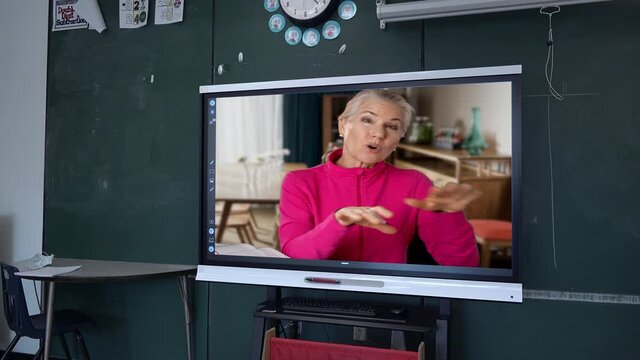 Female teacher gives a grammar lesson on an interactive whiteboard at the front of a classroom set against a blackboard in a school. Remote teaching learning concept.