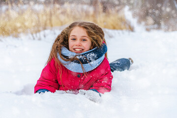 Fototapeta na wymiar A charming smiling little girl with gorgeous hair lies in the snow and laughs